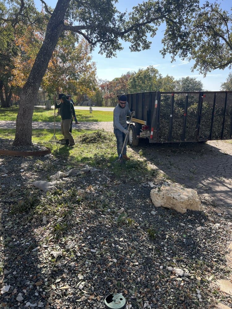 Landscapers raking leaves and branches from a gravel area into a black utility trailer.