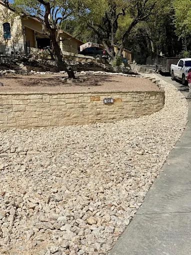 Tan stone retaining wall behind a large bed of light gravel next to a concrete sidewalk.